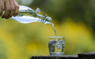 mão derramando beber água da garrafa em vidro com fundo natural foto