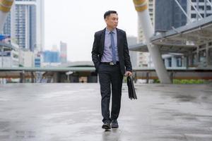 empresário asiático andando e segurando uma pasta com prédios de escritórios comerciais ao fundo da cidade foto