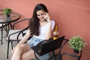 uma jovem mulher é sentado em a terraço com uma computador portátil e uma telefone foto