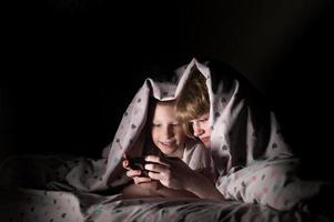 irmão e irmã mentira debaixo uma cobertor às noite e jogar em a telefone foto