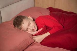 fofa Garoto dentro vermelho pijamas dormindo com dele olhos fechadas dentro cama e sorridente foto