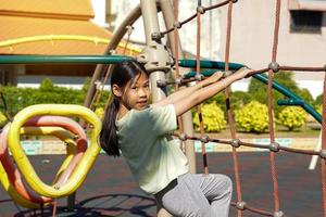menina asiática jogando rede de corda de escalada no playground. playground de conceito, desenvolvimento infantil, esportes e recreação. foco suave e seletivo. foto