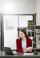 jovem mulher asiática atraente sorrindo pensando planejando escrever em notebook, tablet e laptop trabalhando no escritório foto