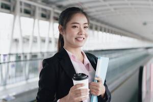 jovem empresária asiática de terno preto segurando a pasta e a xícara de café e de pé na estação de passarela. foto