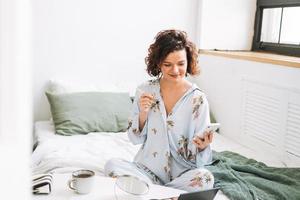 jovem morena mulher dentro azul pijamas aplica-se Maquiagem com Móvel telefone sentado em cama às casa foto