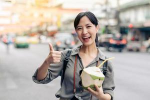 feliz jovem ásia mulher mochila viajante bebendo uma coco suco às China Cidade rua Comida mercado dentro Bangkok, tailândia. viajante verificação Fora lado ruas. foto