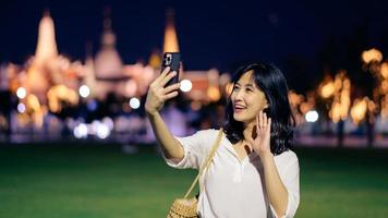 retrato jovem lindo ásia mulher sorridente e usando Smartphone enquanto viagem às têmpora do a esmeralda Buda ou wat phra kaew dentro noite Visão apontar, Bangkok, tailândia. foto