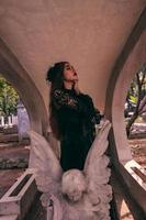uma jovem bruxa dentro uma Preto vestir é em pé entre a lápides em forma gostar a anjo enquanto visitando a cemitério foto