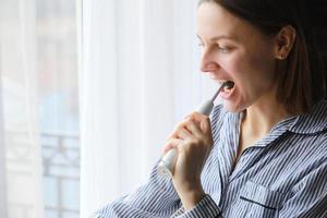 fechar acima em mulheres escovar dentes com elétrico escova de dente de a janela às casa dentro banheiro. cuidados de saúde foto