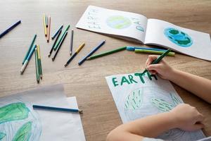 uma criança às escola às a mesa desenha a planeta terra com uma mundo mapa com multicolorido lápis e ponta de feltro canetas em branco papel. Paz e terra dia conceito. foto