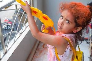 doce pequeno indiano menina jogando cores em holi festival, segurando pichakaree cheio do cores, holi festival celebrações dentro Délhi, Índia foto