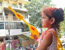 doce pequeno indiano menina jogando cores em holi festival, segurando pichakaree cheio do cores, holi festival celebrações dentro Délhi, Índia foto