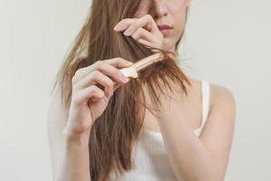 estragado cabelo, frustrado ásia jovem mulher, menina mão dentro segurando escova divisão termina bagunçado enquanto pentear cabelo, não escovado seco grandes cabelo. saúde Cuidado beleza, retrato isolado em branco fundo. foto