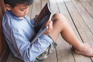 menino sentado contra a porta de madeira lendo um livro no chão de madeira foto