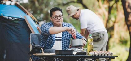 ásia homem preparando arremesso uma barraca dentro acampamento enquanto a de outros homem estava preparando Comida foto