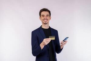 retrato do feliz jovem homem de negocios em pé usando Móvel célula telefone e segurando crédito banco cartão isolado em branco cor fundo estúdio foto