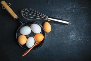 Ovos crus em uma frigideira com colher de pau e bata no fundo de uma mesa de madeira foto