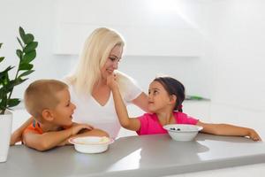 família tendo café da manhã dentro uma branco ensolarado cozinha. jovem mãe alimentando dois crianças, comendo fruta e laticínio. saudável nutrição para crianças. pai com criança pequena criança e bebê cozinhando manhã refeição. foto