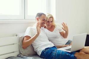 feliz casal falando e acenando mão dentro uma vídeo conferência em linha com uma computador portátil em uma sofá às casa foto