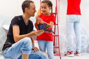 jovem família fazendo uma casa reforma e pintura quartos, a pai é pintura paredes com uma pintura rolo, a mãe e dela filho estão mexendo pintura dentro uma balde foto