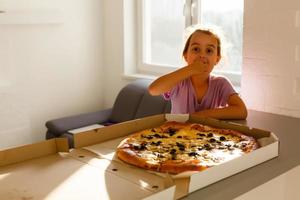 encantador feliz jovem menina rir e morder fora grande fatia do fresco fez pizza. ela sentar às branco cadeira dentro provence estilo interior, sorrir e apreciar ensolarado dia e gostoso refeição. ela tem grandes Loiras cabelo. foto