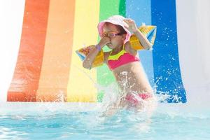 feliz rindo pequeno menina jogando em água deslizar dentro ao ar livre natação piscina em quente verão dia. crianças aprender para nadar. criança vestindo Sol proteção erupção cutânea guarda deslizante em aqua Parque infantil dentro tropical recorrer foto