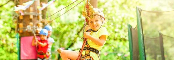 bravo jovem menina dentro capacete sobe em árvore tops dentro diversão corda parque em verão feriados, crianças acampamento foto