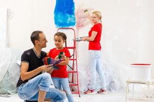 jovem família fazendo uma casa reforma e pintura quartos, a pai é pintura paredes com uma pintura rolo, a mãe e dela filho estão mexendo pintura dentro uma balde foto