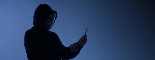 hacker anônimo e máscara facial com smartphone na mão. homem de camisa de capuz preto segurando e usando o celular em fundo branco. represente o hacking de dados de crimes cibernéticos ou o roubo do conceito de dados pessoais foto