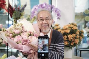 comércio eletrônico negócios, 1 Senior masculino florista trabalhadores demonstrar e mostrar floral arranjos através da conectados viver transmissão com Smartphone inscrição dentro uma brilhante flor comprar, uma lindo flores loja. foto