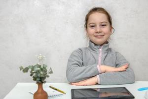 pequeno fofa menina aluna com uma sorrir senta às uma escrivaninha com dela mãos apertado pronto para Aulas, costas para escola foto