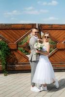 jovem casal noiva dentro uma branco curto vestir e noivo dentro uma cinzento terno perto a de madeira portão foto