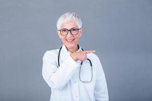 médico Senior mulher, médico profissional apontando dentro certo direção isolado sobre azul fundo. fêmea maduro médico com estetoscópio foto