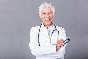 retrato do Senior fêmea médico com estetoscópio braço Cruz isolado em fundo. a médico em pé com alegre gesticulando. ela vestindo médico uniforme dentro hospital. saúde seguro conceito. foto