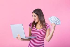 retrato do animado satisfeito menina segurando dinheiro notas com computador portátil computador isolado sobre Rosa fundo. retrato do uma alegre jovem mulher segurando dinheiro notas e computador portátil computador dentro mãos foto