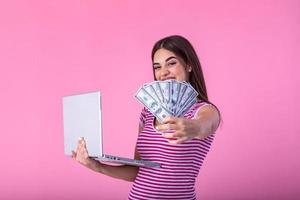 retrato do a animado satisfeito menina segurando dinheiro notas com computador portátil computador isolado sobre Rosa fundo. retrato do uma alegre jovem mulher segurando dinheiro notas e notebook dentro mãos foto