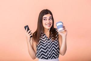 retrato do alegre juventude segurando quente bebida olhando morder lábio e segurando esperto Móvel telefone isolado sobre pêssego fundo. sorridente menina dentro com levar longe café copo e célula telefone . foto