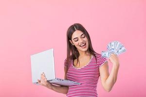 imagem do alegre jovem mulher em pé isolado sobre Rosa fundo usando computador portátil computador e segurando dinheiro notas . retrato do uma sorridente menina segurando computador portátil computador foto