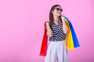 jovem atraente menina dentro oculos de sol desfrutando uma Boa compras , com bolsas dentro mão. jovem feliz verão compras mulher com compras bolsas isolado em Rosa fundo foto