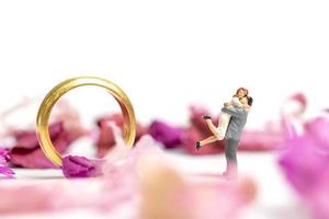 casal em miniatura se abraçando com uma aliança de casamento e pétalas de rosa isoladas em um fundo branco foto