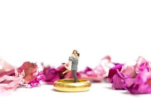 casal em miniatura se abraçando com uma aliança de casamento e pétalas de rosa isoladas em um fundo branco foto