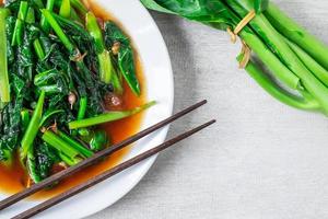 Couve chinesa frita com molho de ostra com pauzinhos em um prato branco ao lado de couve chinesa fresca na mesa de madeira foto