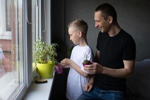 uma fofa Garoto é rega interior plantas. Papai Próximo para dele filho perto a janela foto