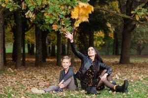 uma menina e uma Garoto estão sentado em a terra e jogando outono folhas foto
