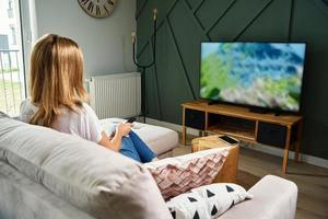 mulher Assistir televisão conjunto em a sofá e interruptor canais com controlo remoto ao controle foto