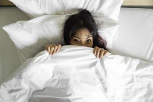 uma engraçado ásia mulher deitado dentro cama e se escondendo debaixo uma branco cobertor, olhando às a Câmera com olhos cheio do alegria. foto