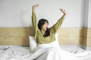 fofa ásia mulher alongamento Fora e bocejando enquanto sentado em dela cama dentro brilhante quarto foto