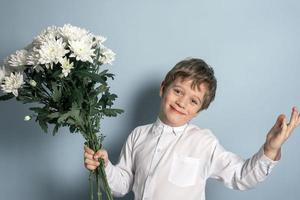 uma fofa caucasiano Garoto dentro uma branco camisa detém uma ramalhete do branco flores dentro dele mão e sorri. foto