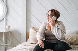 sorridente meio envelhecido mais Tamanho mulher usando Móvel telefone em cama às casa foto