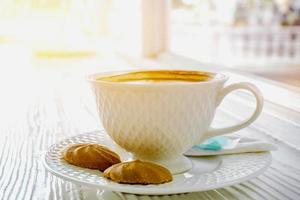 quente café dentro uma branco café copo com biscoitos em branco de madeira mesa em Sol flare fundo. foto